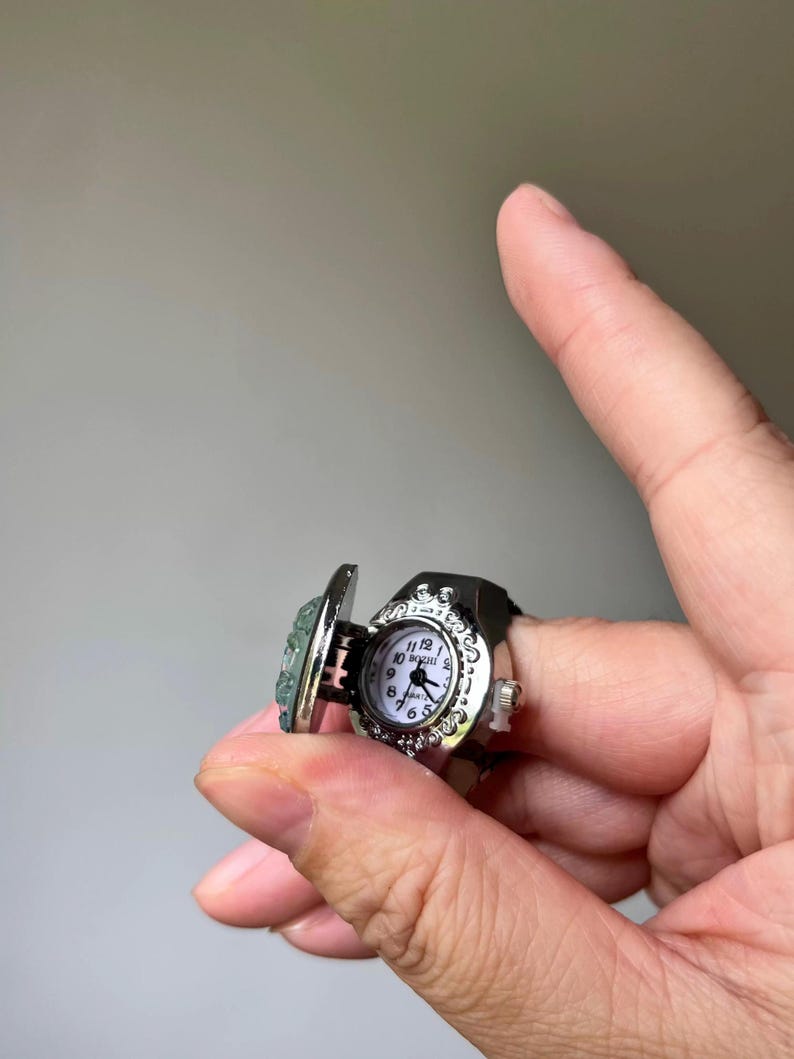 May include: A silver-toned ring watch with a hinged cover. The watch face is white with black numerals and hands. The ring has a decorative bezel and a black band. The hinged cover has a light blue stone.