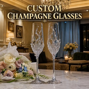 May include: Two clear glass champagne flutes decorated with intricate silver and white crystal embellishments on the bowls and bases. The text CUSTOM CHAMPAGNE GLASSES is displayed in gold lettering above the glasses.