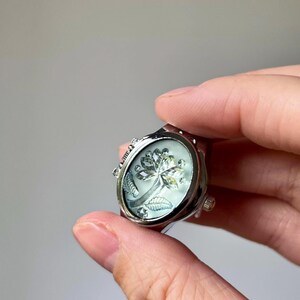 May include: Silver-toned ring with an oval face featuring a floral design. The ring has a detailed, ornate setting and a light-colored, possibly mother-of-pearl, background. The floral design appears to be a stylized flower with leaves.