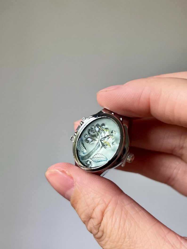 May include: Silver-toned ring with an oval face featuring a floral design. The ring has a detailed, ornate setting and a light-colored, possibly mother-of-pearl, background. The floral design appears to be a stylized flower with leaves.