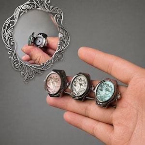 Peut inclure: Trois bagues de style antique avec des motifs floraux roses, blancs et turquoise sont présentées sur une main. Une bague montre miniature est reflétée dans un miroir décoratif. Les bagues ont une finition en métal foncé.