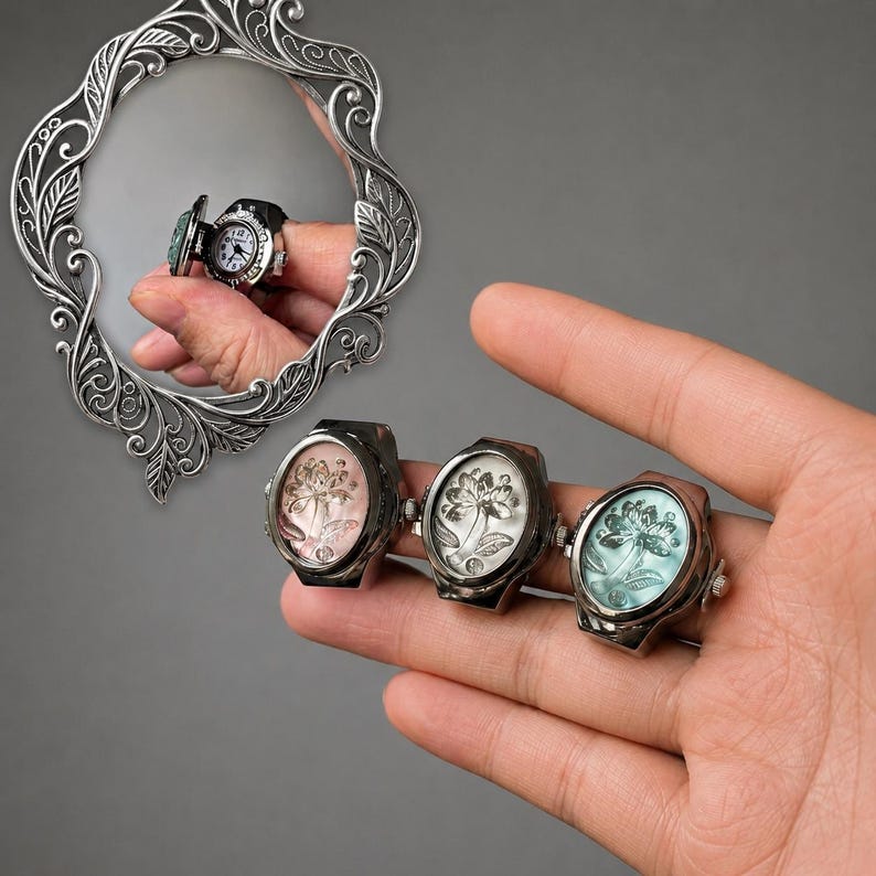 May include: A hand displays three antique-style rings with floral designs in pink, white, and teal. A fourth ring, a miniature watch, is reflected in a decorative mirror. The rings have a dark metal finish.