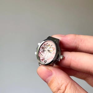 May include: Silver-toned ring with an oval face featuring a floral design on a pink background. The ring has a watch-like appearance with small dials on the sides. The floral design is detailed with gold and silver accents.