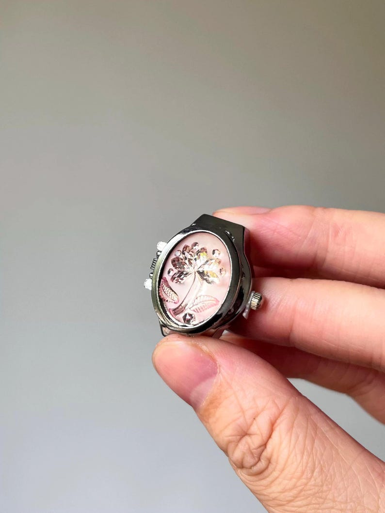 May include: Silver-toned ring with an oval face featuring a floral design on a pink background. The ring has a watch-like appearance with small dials on the sides. The floral design is detailed with gold and silver accents.