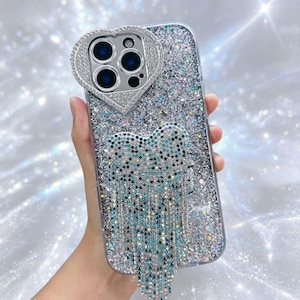 Puede incluir: Una funda de teléfono transparente con acabado de purpurina plateada y un marco de cámara en forma de corazón incrustado con pedrería. Un gran lazo colgante hecho de cristales azules y plateados cuelga del centro de la parte trasera.