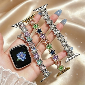 Pode incluir: Um smartwatch prateado com um mostrador de flor azul e três pulseiras decorativas. Cada pulseira apresenta uma série de cristais conectados em forma de estrela em várias cores, incluindo rosa, roxo, verde, azul e transparente.