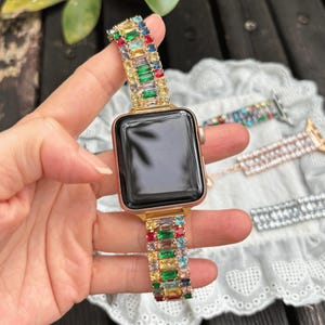 Könnte beinhalten: Eine goldfarbene Smartwatch mit schwarzem Display und einem dekorativen Uhrenarmband, das mit rechteckigen, mehrfarbigen Baguette-Kristallen in Rot, Grün, Blau und Gelb besetzt ist, die in einem sich wiederholenden Muster angeordnet sind.