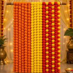 Op de afbeelding: Een decoratieve wandhanger met verticale strengen kunstmatige goudsbloemen in levendige tinten oranje, geel en rood. De strengen hangen aan een horizontale houten roede, wat een dicht, kleurrijk gordijn van bloemige pompons creëert.
