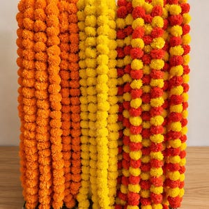 Puede incluir: Primer plano de varias hebras de guirnaldas de caléndulas en tonos naranja, amarillo y rojo. Las guirnaldas están hechas de pequeñas flores redondas y están dispuestas verticalmente. El fondo es de color claro.