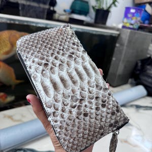 May include: A rectangular wallet featuring a textured snakeskin pattern in shades of white, grey, and dark brown. The wallet has a black zip closure along the edge and a matching snakeskin zip pull tab attached to the metal slider mechanism.
