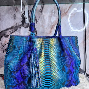 May include: A blue and gold snakeskin-patterned tote bag with a tassel. The bag has a structured shape with a top handle and a shoulder strap. The bag's colors range from deep blue to turquoise and gold.