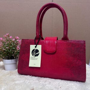 Puede incluir: Un bolso fucsia vibrante con un patrón texturizado similar al de un reptil. El bolso tiene dos asas redondeadas y un cierre de solapa. Una etiqueta con la marca "ANNIDAP" cuelga de un asa. El bolso está sobre una superficie blanca y esponjosa.