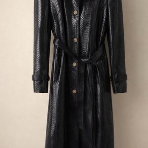 May include: A long, black trench coat with a textured, snakeskin-like pattern. The coat has a belted waist, gold-coloured buttons, and a red lining. The sleeves have buttoned cuffs, and the coat is knee-length.
