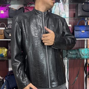 May include: A black leather jacket with a snakeskin pattern. The jacket features a zip closure and a stand-up collar. The jacket is displayed in a shop with various handbags on shelves in the background.