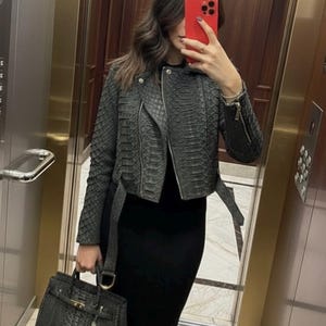 May include: A dark gray, textured leather jacket with a zipper closure and belt, paired with a black dress and a matching handbag. The handbag has a top handle and a shoulder strap.
