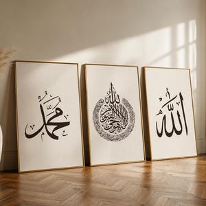 May include: Three framed art prints with gold frames. Each print features black Arabic calligraphy on a cream background. The prints are leaning against a wall on a wooden floor. A vase with dried plants is to the left.