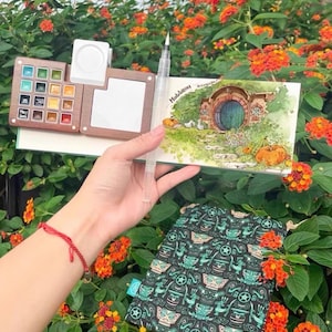 Puede incluir: Un kit de pintura de acuarela con una paleta de madera, un pincel de agua y un cuaderno de bocetos con una ilustración de Hobbiton. También se ve una bolsa estampada en negro y verde azulado. La escena está ambientada al aire libre con flores naranjas vibrantes.