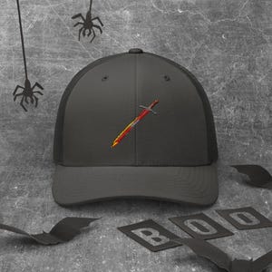 Può includere: Cappellino da camionista grigio scuro con retro in rete e visiera curva. Il cappellino presenta un disegno di spada ricamata in rosso, giallo e arancione. La parola "BOO" è scritta con lettere ritagliate.
