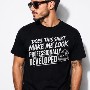 May include: A black short-sleeved t-shirt featuring white text that reads DOES THIS SHIRT MAKE ME LOOK PROFESSIONALLY DEVELOPED along with a graphic of a laptop displaying code brackets and a gear icon on the right side of the chest area.
