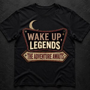 May include: A black short-sleeve t-shirt featuring a graphic design of a vintage-style sign with the text "WAKE UP, LEGENDS" and "THE ADVENTURE AWAITS" in cream and red lettering, accented by a crescent moon and small star icons above the sign.