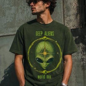 May include: A dark olive green short-sleeve t-shirt featuring a graphic of a green alien head centered within a yellow circular frame. The text "DEEP ALIENS" is printed above the graphic, and the text "world mix" is printed below the graphic.