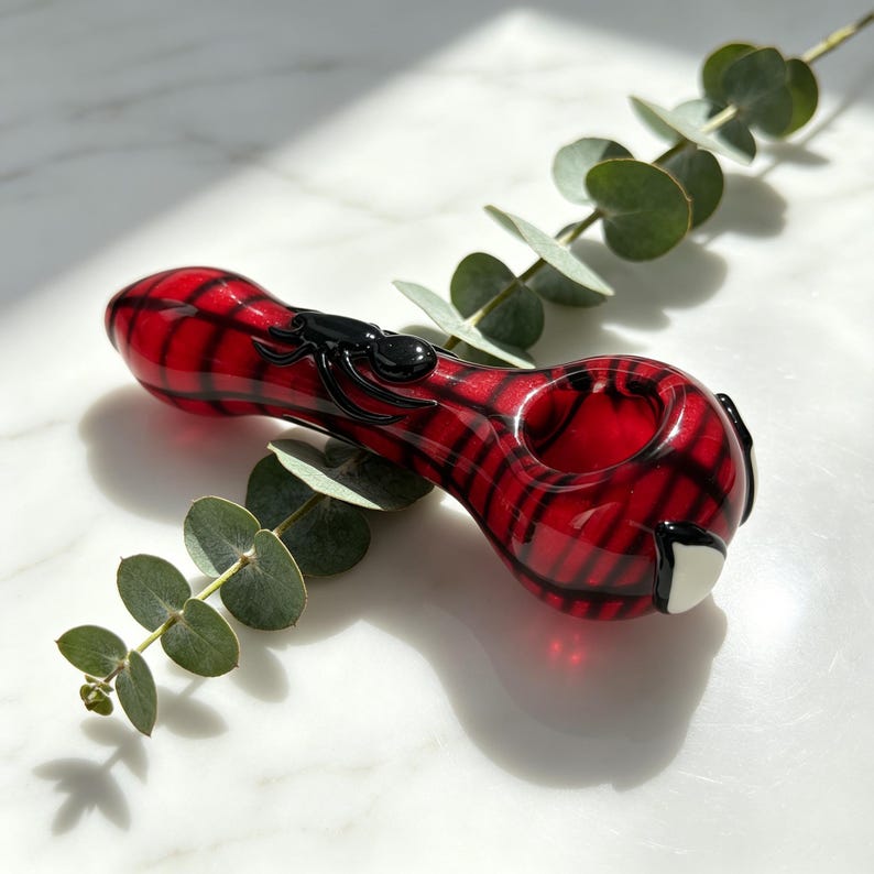 May include: A red glass pipe featuring a black spider design and black grid-like patterns. The pipe has a rounded bowl and a tapered stem, with a small white accent detail near the bowl. The glass is translucent and smooth in texture.