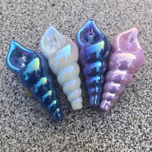 May include: Four iridescent seashell ornaments in shades of blue, white, purple, and pink. The glossy shells have a spiral design and a pearlescent sheen, reflecting light with a rainbow effect. These decorative items are ideal for coastal or nautical themed decor.