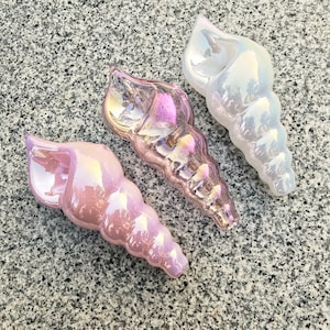 Pode incluir: Três ornamentos de conchas iridescentes em tons de rosa e branco. As conchas decorativas têm um acabamento perolado, refletindo a luz com um efeito arco-íris. As conchas estão dispostas sobre uma superfície cinza salpicada, mostrando suas formas e cores únicas.