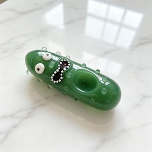 May include: A green glass pipe shaped like a pickle with a textured surface featuring small bumps. The pipe has a cartoon-like face with two white eyes and a wide, open mouth with white teeth, and a circular bowl integrated into the body.