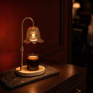 Puede incluir: Una lámpara calentadora de velas de metal dorado con un brazo curvo y una pantalla de vidrio estriado en forma de campana. La lámpara se asienta sobre una base de madera clara que sostiene un frasco de vela de vidrio oscuro con luz cálida.