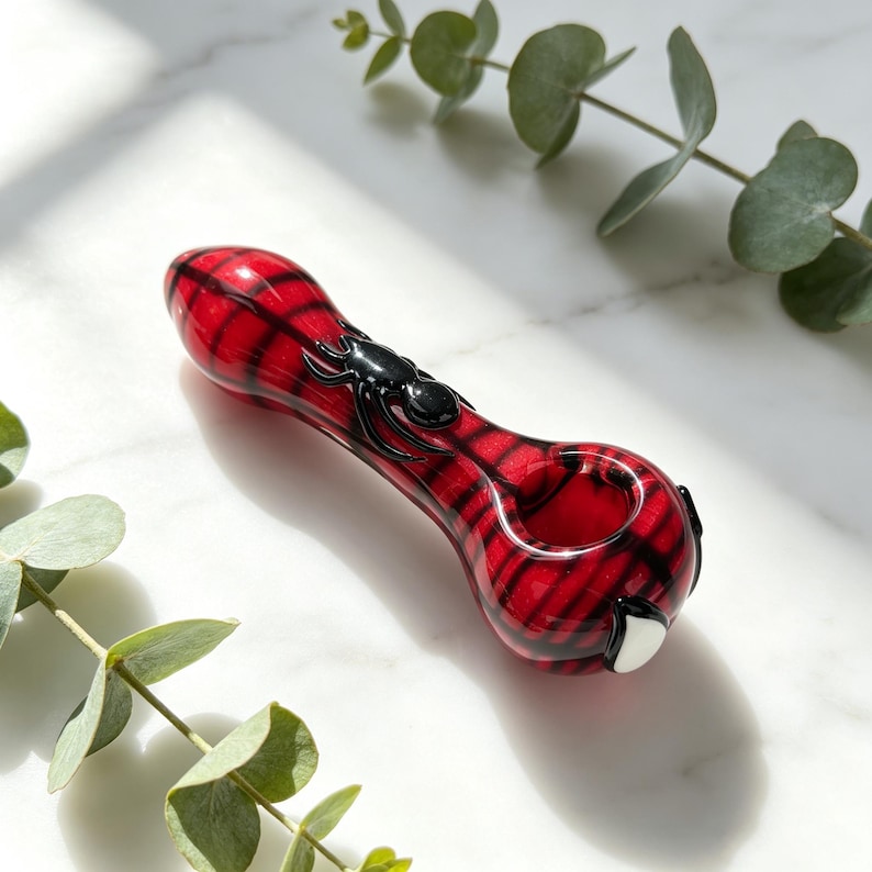 May include: A red glass pipe featuring a black spider design and a dark web pattern across the surface. The pipe has a rounded bowl and a smooth, glossy finish, with a small white accent detail near the base of the bowl.
