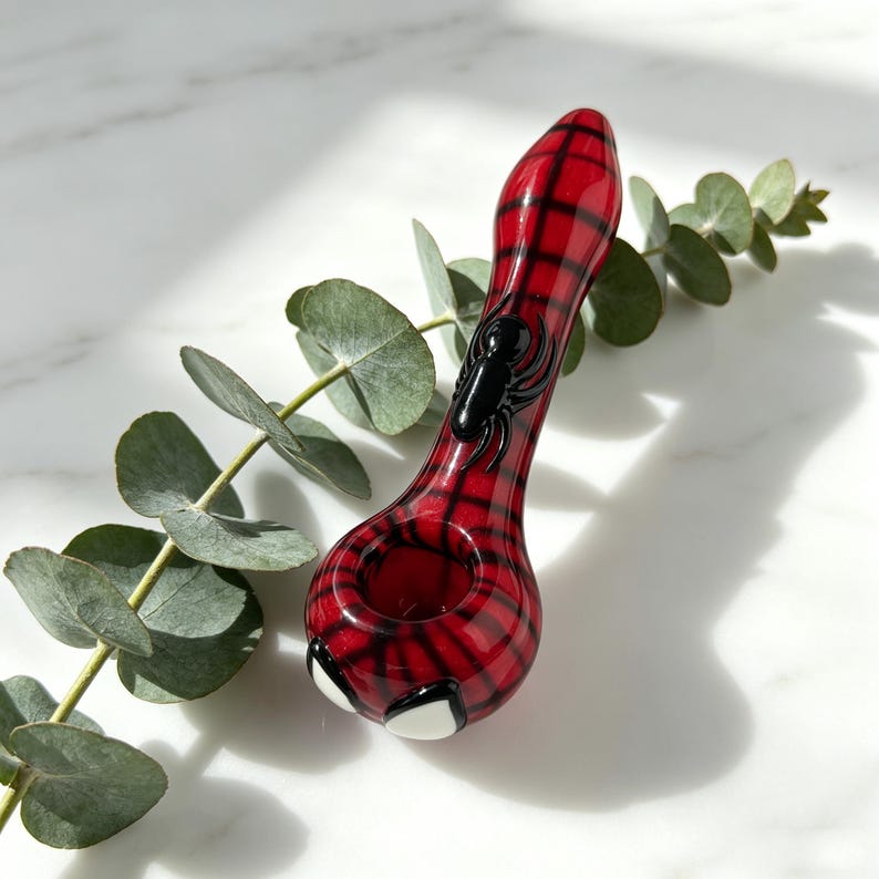 May include: A glass pipe featuring a red and black plaid pattern, a raised black spider emblem on the stem, and two white eye-shaped accents near the bowl. The pipe has a smooth, glossy finish and a rounded shape, showcasing intricate glasswork.