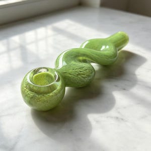 May include: A light green glass pipe with a wavy, serpentine body and a circular bowl at one end. The glass features a translucent, marbled texture throughout, with a smooth, glossy finish that reflects light across its curved, elongated form.