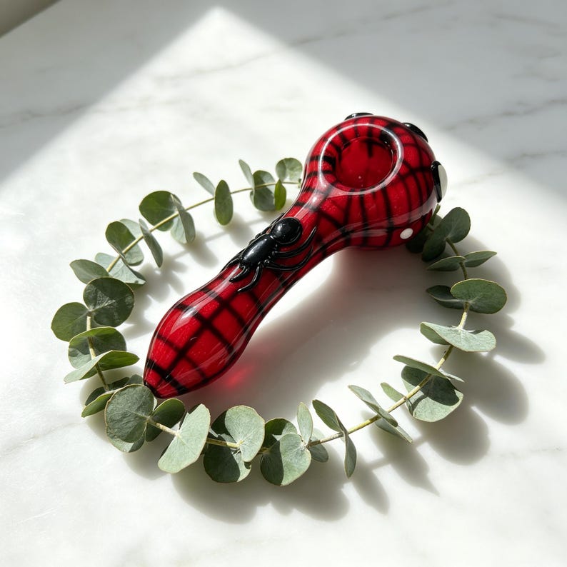 May include: A glass pipe featuring a vibrant red and black plaid pattern. The pipe has a smooth, rounded bowl and a tapered stem, adorned with a small, three-dimensional black spider figure attached to the top surface of the glass stem.