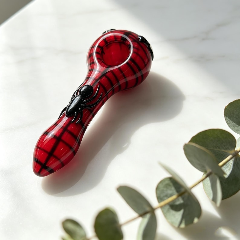 May include: A glass pipe featuring a vibrant red base with a black grid pattern resembling a spider web. A small, three-dimensional black spider figure is attached to the stem of the pipe, which has a rounded bowl at the top end.
