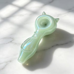 May include: A pale mint green glass pipe featuring an iridescent finish and a cat-shaped bowl with two small pointed ears at the top. The stem includes a raised, curved glass detail resembling a tail, with a smooth, glossy, and translucent surface.