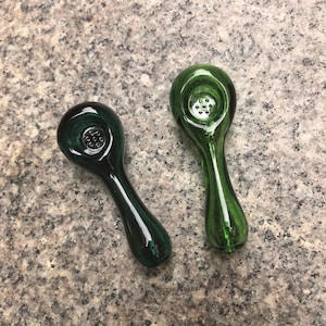 May include: Two glass pipes with smooth, rounded bowls and tapered stems. One pipe is a deep, dark forest green, while the other is a lighter, translucent emerald green. Each bowl features a small, circular filter screen with multiple holes.