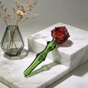 May include: A glass pipe in the shape of a rose. The bowl is a deep red, and the stem is green. The pipe is on a white marble surface. A geometric glass vase with dried flowers is in the background.
