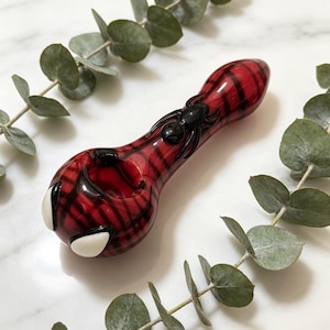 Op de afbeelding: Een glazen pijp met een rood en zwart ruitpatroon, versierd met een zwart spinnenfiguurtje op de steel en twee witte, amandelvormige accenten bij de kop. De pijp heeft een gladde, glanzende afwerking en een afgeronde, bolvormige vorm.