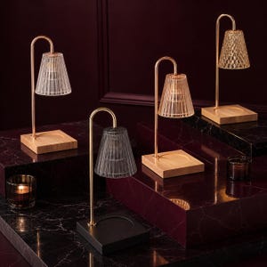 Puede incluir: Cuatro lámparas calentadoras de velas con brazos de metal en tono dorado y pantallas de vidrio estriado. Tres lámparas tienen bases de madera clara, mientras que una tiene una base negra. Cada base incluye una hendidura circular.