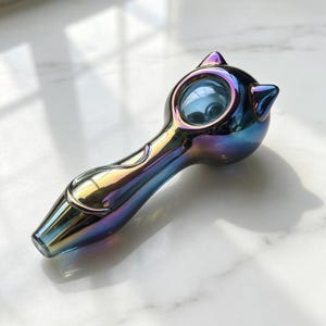 May include: A glass smoking pipe with an iridescent, multi-colored finish that shifts between blue, purple, and gold tones. The bowl features two small, pointed ears at the top, and a curved, tail-like design embossed along the handle.