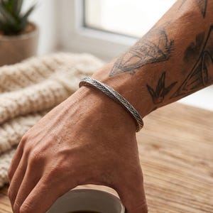 May include: A silver cuff bracelet featuring a detailed, woven herringbone pattern across its entire surface. The metal has a polished finish with darkened recessed areas that highlight the intricate braided texture of the band design.