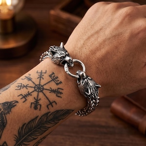 May include: A silver-toned metal bracelet featuring two detailed wolf head terminals connected by a circular ring clasp. The bracelet is constructed with a woven chain link design, showcasing a metallic finish with dark oxidized accents in the crevices.