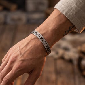 Puede incluir: Una pulsera de brazalete de metal en tono plateado que presenta una serie de caracteres rúnicos grabados a lo largo de la superficie exterior. La pulsera tiene un acabado mate y un diseño simple y abierto que descansa sobre la muñeca.