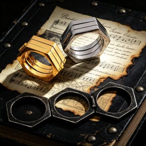 May include: Two hexagonal rings, one gold and one silver, with a layered design. The rings sit on a vintage book with sheet music. Below are three open, dark metal hexagonal rings.