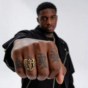 May include: A gold-colored ring with an eagle and sword design. The ring is worn on a hand with several tattoos. The ring's design features an eagle with outstretched wings, a sword, and other decorative elements.