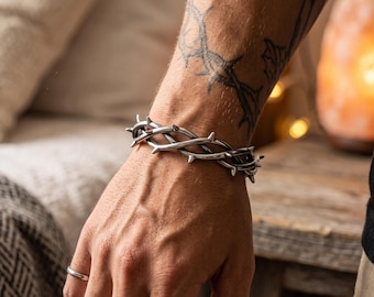 Può includere: Un bracciale in metallo color argento progettato con un motivo a vite intrecciata caratterizzato da spine affilate e sporgenti. Il bracciale circonda il polso, mostrando una finitura metallica lucida con dettagli intricati che imitano l'aspetto di una corona di spine.