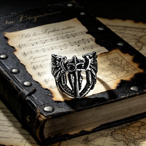 May include: A silver-colored ring with a detailed design featuring a sword, wings, and other ornate elements. The ring is set against a backdrop of an antique book and aged paper with musical notes.
