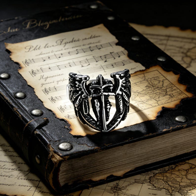 May include: A silver-colored ring with a detailed design featuring a sword, wings, and other ornate elements. The ring is set against a backdrop of an antique book and aged paper with musical notes.