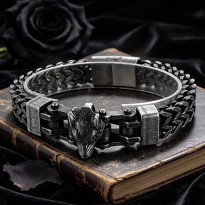 May include: A dark silver-toned bracelet featuring a central wolf head motif integrated into a mechanical chain-link design. The bracelet has a weathered, distressed metal finish and a rectangular clasp mechanism, showcasing a rugged aesthetic.